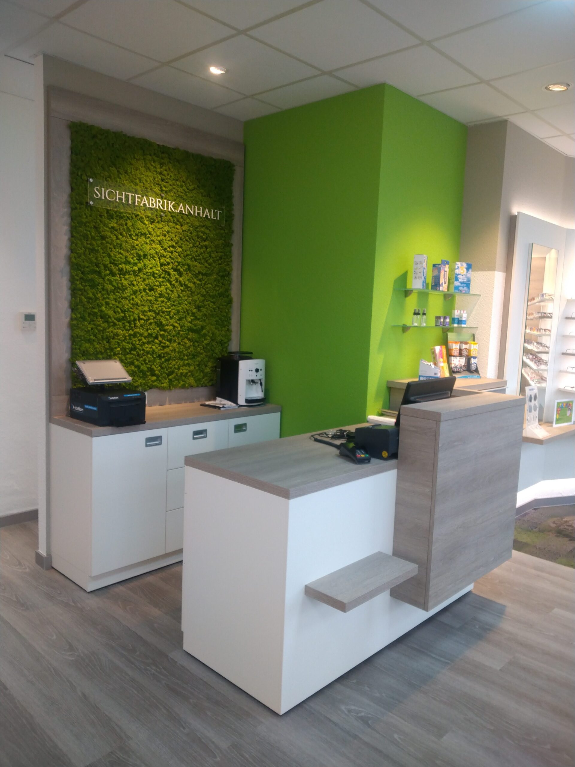 Modernes Empfangsbereich mit grüner Wand, Theke, Kaffeestation und Brillenregal.